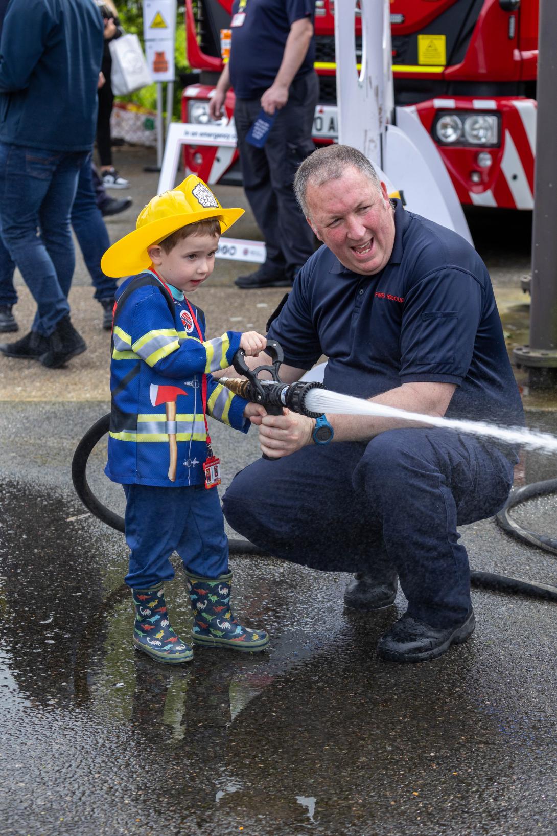 A child, dressed as a firefighter, gets instructed on how to use the fire hose